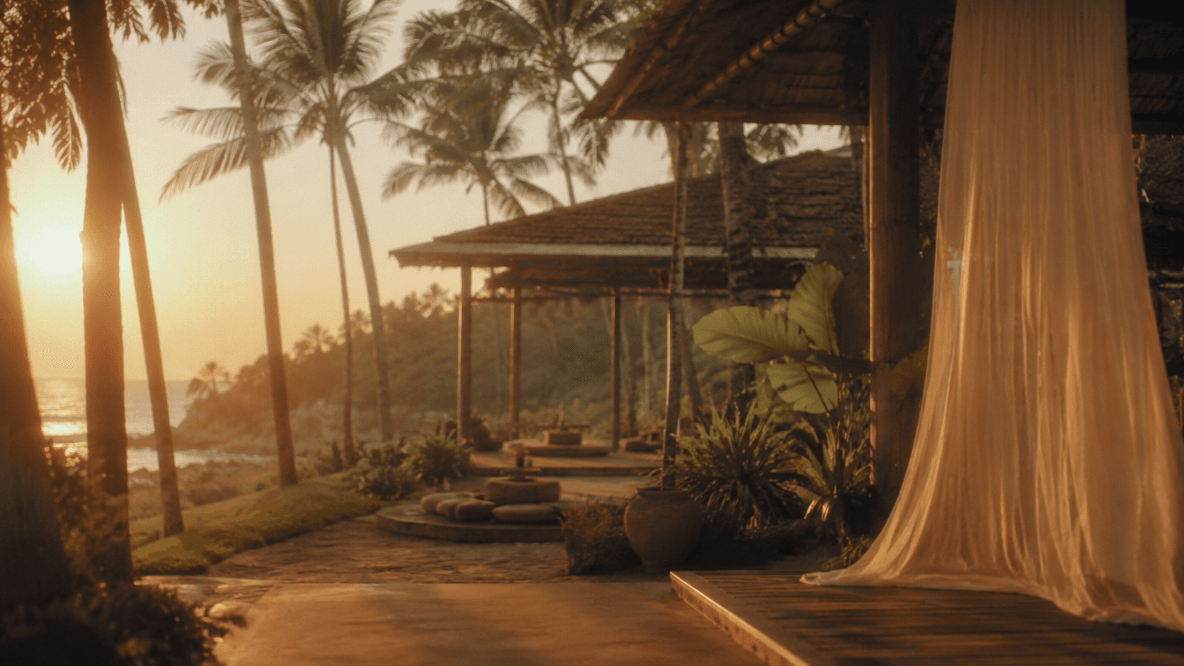 An open-air pavilion with sheer curtains at sunset, palms and the sea beyond